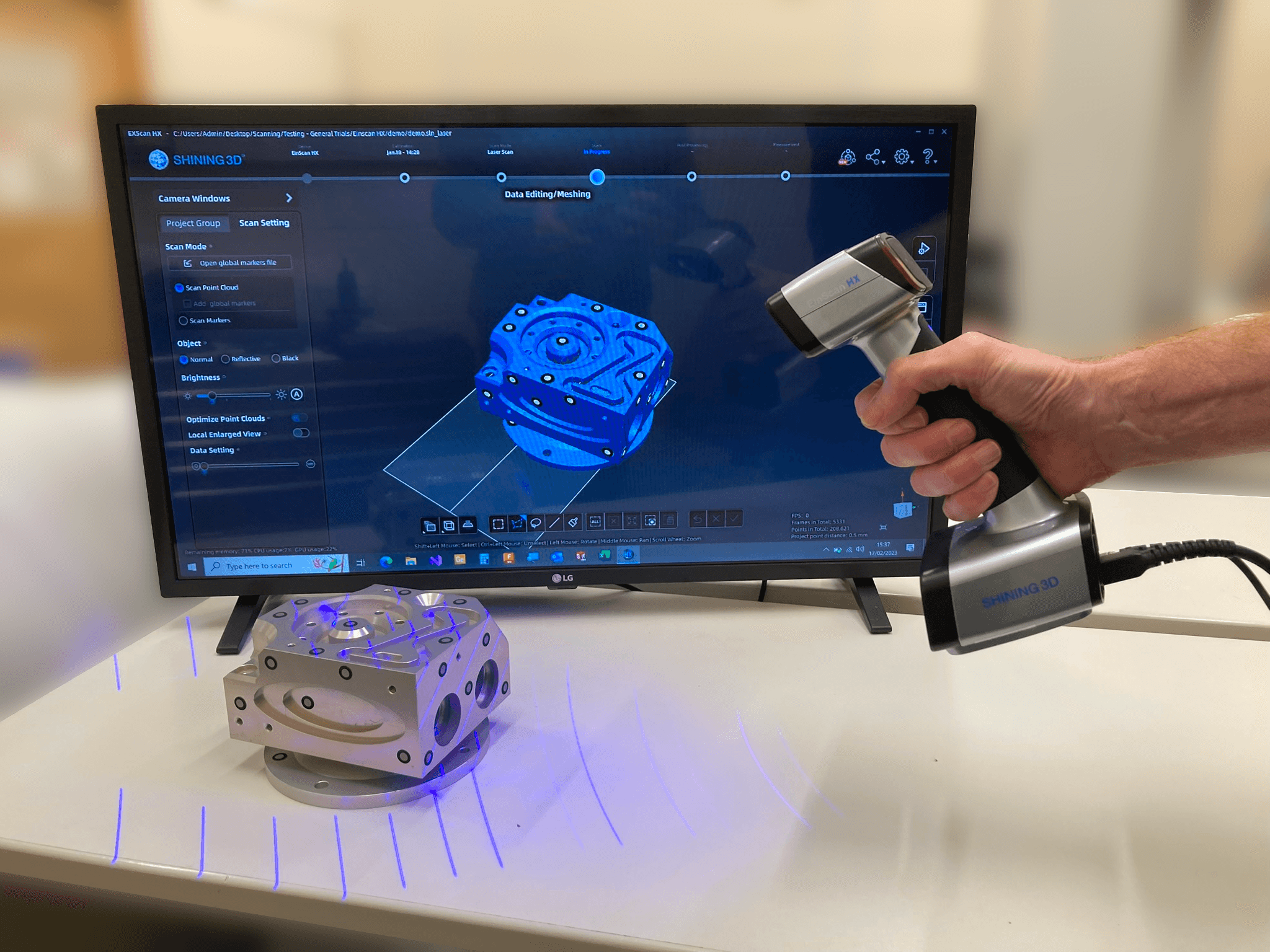 A handheld 3D scanner pointing at a small detailed part. A screen shows the captured scan data.